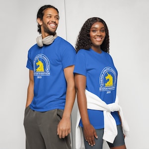 May include: Two people wearing royal blue t-shirts with a white circle graphic featuring a yellow unicorn and the text "Boston Athletic Association Boston Marathon 2025".