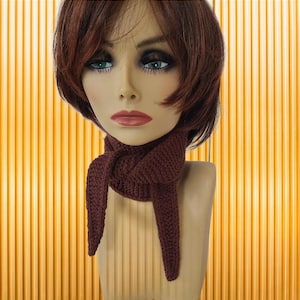 May include: A burgundy-colored knitted scarf is draped around a mannequin's neck. The scarf is knotted at the neck and has two long, pointed ends. The mannequin has brown hair, blue eyes, and red lipstick. The background is a vertical striped pattern in shades of yellow and orange.
