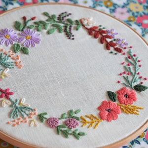 Può includere: Ricamo floreale fatto a mano con una ghirlanda di fiori colorati. Il design include margherite viola, papaveri rossi e altri fiori in tonalità di rosa, giallo e verde, il tutto su un tessuto color crema.