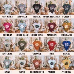 May include: A collection of 20 t-shirts in various colours, each featuring the text "Your Design Here" on the front. The t-shirts are displayed on hangers against a backdrop of woven wicker.