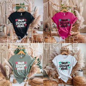 May include: Four t-shirts in different colours, each featuring the text "Your Design Here" and hearts printed on the front. The shirts are displayed on a wooden hanger and are hanging in front of a backdrop of woven baskets and dried pampas grass.