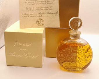 Puede incluir: Una caja dorada con el texto "Baccarat pour Annick Goutal" en la parte delantera. Una botella de vidrio transparente con un tapón dorado y un diseño dorado en la botella. La botella está sentada sobre una caja dorada con un certificado de autenticidad para el perfume.