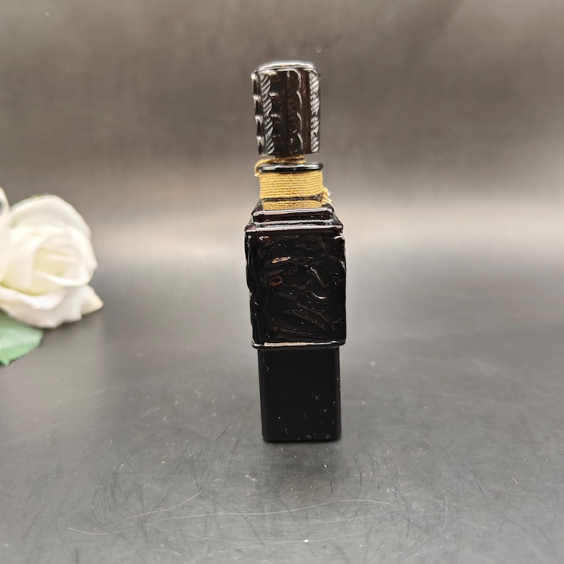 May include: A black bottle stopper with a rectangular body and a cylindrical top. The body has carved floral designs, and the top has a textured pattern. The stopper is wrapped with a gold-colored twine.