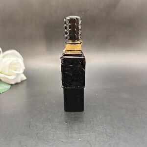 May include: A black perfume bottle with a decorative carved design. The bottle has a rectangular shape with a detailed stopper. The stopper is black with a white design. The bottle is wrapped with gold-colored thread.