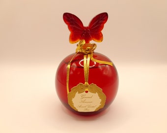 May include: A red glass bottle with a gold butterfly stopper and a gold label that reads "Grand Amour Armand Grantel Paris".