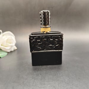 May include: A black perfume bottle with a rectangular base and a decorative top. The bottle features intricate relief carvings and a silver-colored band. The stopper is cylindrical with a textured design and a gold-colored string.