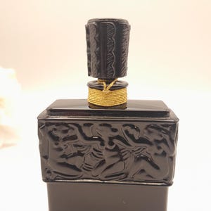 May include: A black perfume bottle with a decorative stopper. The bottle has a rectangular base with a carved relief design. The stopper is cylindrical and has a carved pattern. A gold-colored cord wraps around the base of the stopper.