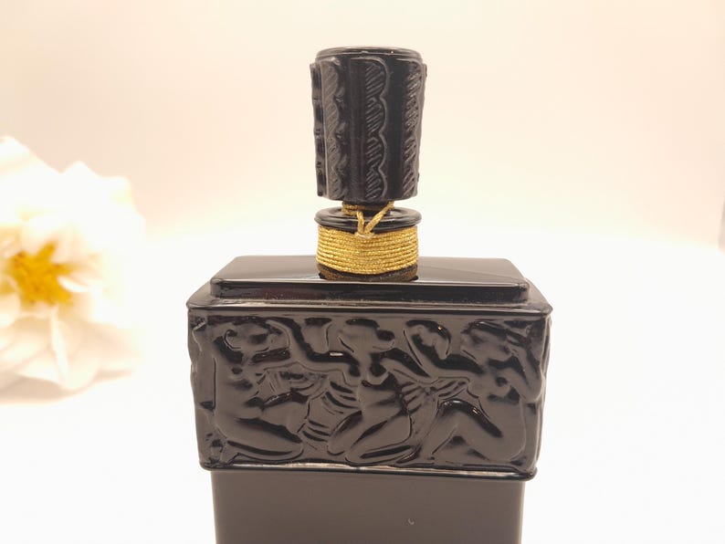 May include: A black perfume bottle with a decorative stopper. The bottle has a rectangular base with a carved relief design. The stopper is cylindrical and has a carved pattern. A gold-colored cord wraps around the base of the stopper.