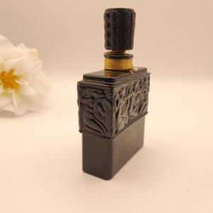 May include: A black rectangular perfume bottle with a decorative stopper. The bottle has a detailed relief design on the front and sides. The stopper is cylindrical and also has a carved design. A gold-colored band encircles the bottle's neck.