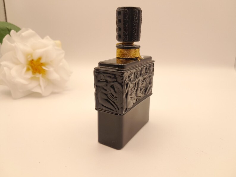 May include: A black rectangular perfume bottle with a decorative stopper. The bottle has a detailed relief design on the front and sides. The stopper is cylindrical and also has a carved design. A gold-colored band encircles the bottle's neck.