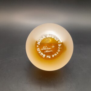 May include: A clear glass bottle with a frosted top and a gold label. The label reads "Nina Ricci. Made in France. Lalique."