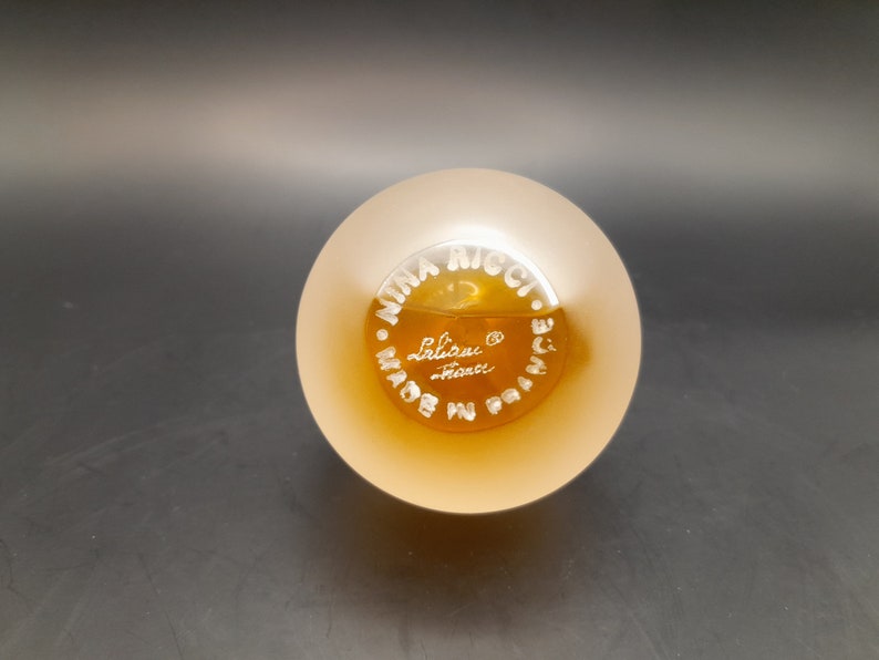 May include: A clear glass bottle with a frosted top and a gold label. The label reads "Nina Ricci. Made in France. Lalique."
