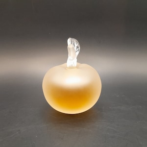 May include: A frosted glass perfume bottle shaped like an apple with a clear glass stopper. The bottle is filled with a light amber liquid.