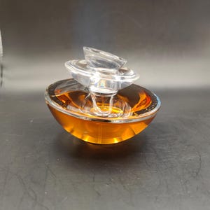 Puede incluir: Un frasco de perfume de vidrio transparente con una tapa de varios niveles. El frasco est&aacute; lleno de un l&iacute;quido &aacute;mbar dorado.