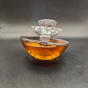 Puede incluir: Un frasco de perfume de vidrio transparente con un dise&ntilde;o abstracto en capas. El frasco est&aacute; lleno de un l&iacute;quido &aacute;mbar dorado.