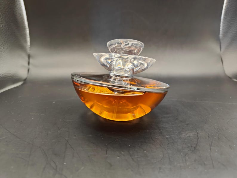 Puede incluir: Un frasco de perfume de vidrio transparente con un dise&ntilde;o abstracto en capas. El frasco est&aacute; lleno de un l&iacute;quido &aacute;mbar dorado.