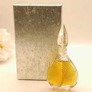 May include: A clear glass perfume bottle with a heart-shaped body and vertical ridges, filled with golden liquid. The bottle has a pointed glass stopper labelled "parfum CHAMADE" and stands in front of a rectangular, silver-textured box.