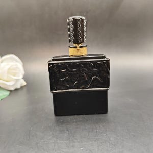 May include: A black perfume bottle with a silver rim and a decorative stopper. The bottle has a rectangular shape with a detailed relief design. The stopper is silver with a textured pattern, and the neck is wrapped with gold thread.
