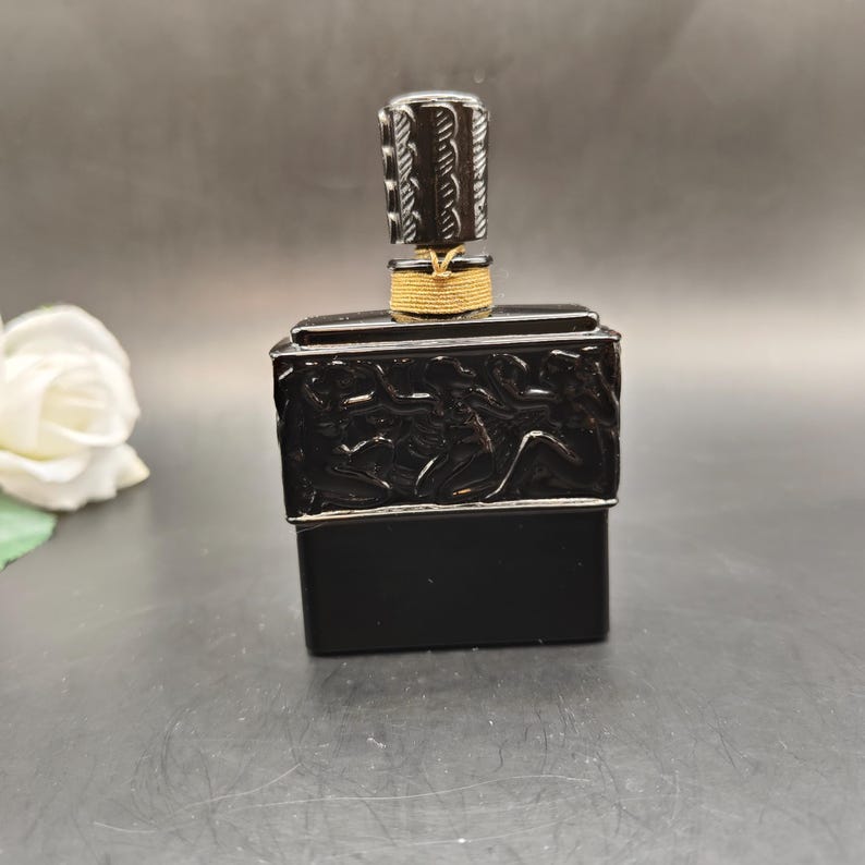 May include: A black perfume bottle with a silver rim and a decorative stopper. The bottle has a rectangular shape with a detailed relief design. The stopper is silver with a textured pattern, and the neck is wrapped with gold thread.