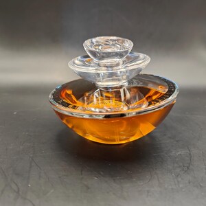 Puede incluir: Un frasco de perfume de vidrio transparente con un dise&ntilde;o en capas y un l&iacute;quido de color dorado en su interior. El frasco tiene una base redonda y un borde plateado.