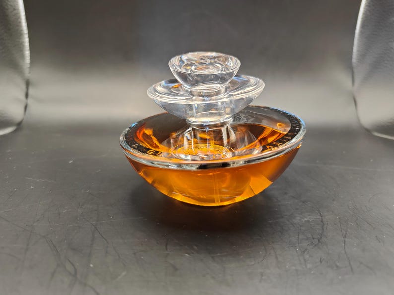 Puede incluir: Un frasco de perfume de vidrio transparente con un dise&ntilde;o en capas y un l&iacute;quido de color dorado en su interior. El frasco tiene una base redonda y un borde plateado.