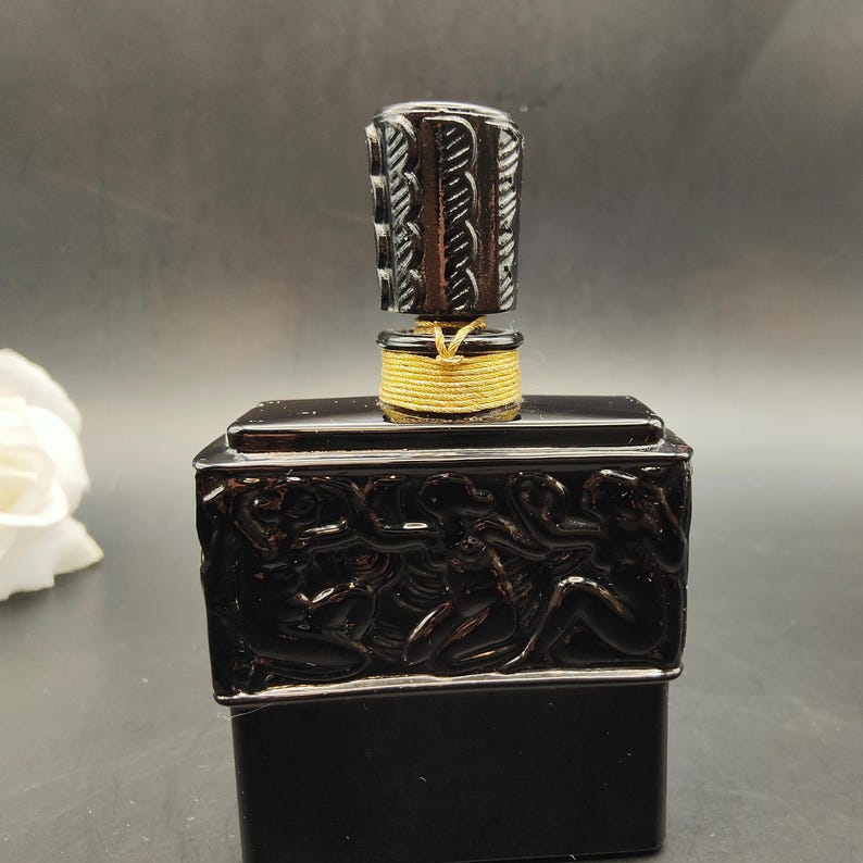 May include: A black perfume bottle with a carved design of figures on the front. The stopper is dark with a rope-like design and a gold-colored string around the base. The bottle is rectangular and sits on a dark surface.