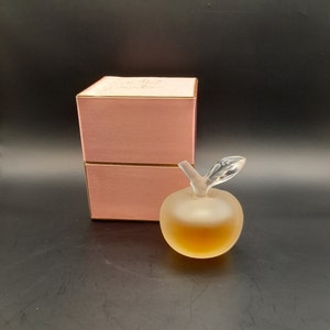 May include: A frosted glass apple-shaped perfume bottle with a gold lid and a pink box. The bottle is filled with a light amber liquid.