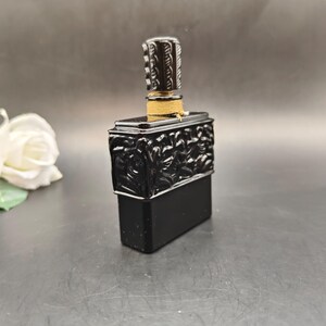 May include: A black rectangular perfume bottle with a decorative top and stopper. The bottle has a detailed relief design on the front and is accented with gold-colored twine around the neck. The stopper is cylindrical with a textured pattern.