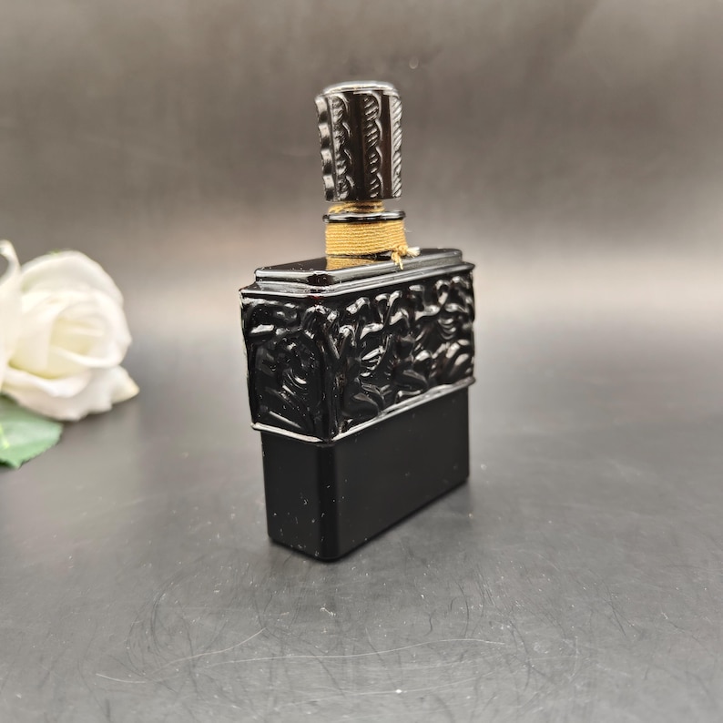 May include: A black rectangular perfume bottle with a decorative top and stopper. The bottle has a detailed relief design on the front and is accented with gold-colored twine around the neck. The stopper is cylindrical with a textured pattern.