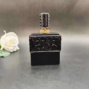 May include: A dark brown perfume bottle with a decorative stopper. The bottle has a rectangular base and a detailed relief design around the top. The stopper is cylindrical with a carved pattern and is secured with a gold-colored twine.