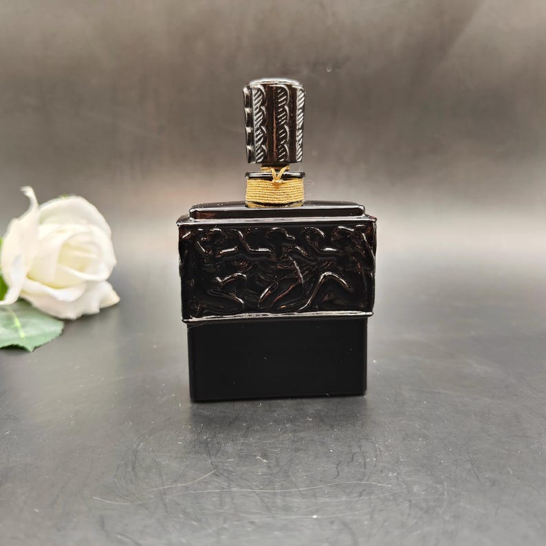 May include: A dark brown perfume bottle with a decorative stopper. The bottle has a rectangular base and a detailed relief design around the top. The stopper is cylindrical with a carved pattern and is secured with a gold-colored twine.