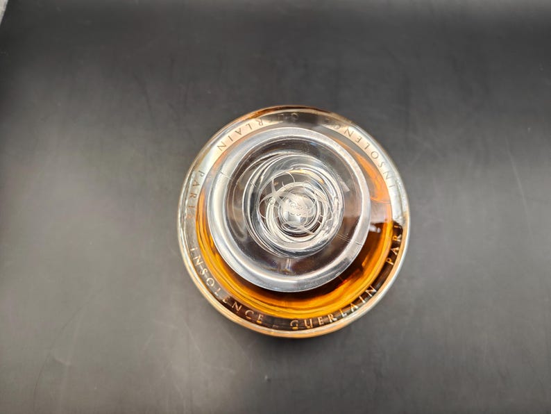 Puede incluir: Una botella de vidrio transparente con una tapa dorada y un l&iacute;quido marr&oacute;n en su interior. La botella est&aacute; etiquetada con "Insolence Guerlain Paris".