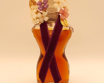 May include: A clear glass perfume bottle with a curved silhouette containing amber liquid. The stopper is adorned with gold-toned metal leaves and small white, purple, and pink flowers. A dark purple velvet ribbon is tied around the bottle neck.