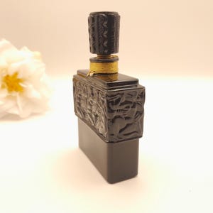 May include: A black rectangular perfume bottle with a decorative stopper. The stopper and the upper part of the bottle have a textured, carved design. The bottle is accented with gold-colored trim. A white flower with a yellow center is in the background.