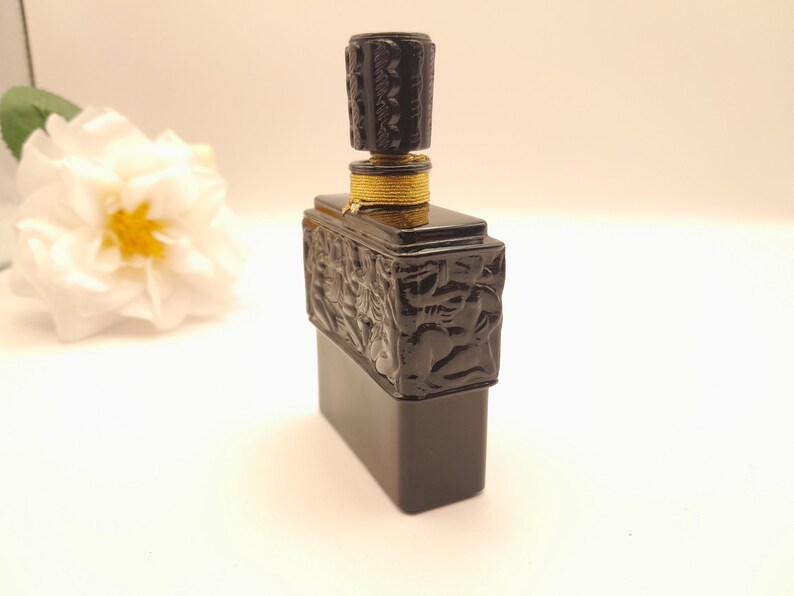 May include: A black rectangular perfume bottle with a decorative stopper. The stopper and the upper part of the bottle have a textured, carved design. The bottle is accented with gold-colored trim. A white flower with a yellow center is in the background.