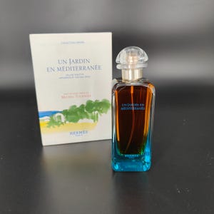 May include: A perfume bottle and box set. The bottle is a gradient of blue to brown with a clear cap. The box is white with the text "UN JARDIN EN MÉDITERRANÉE" and a landscape illustration. The brand name "HERMES" is visible.