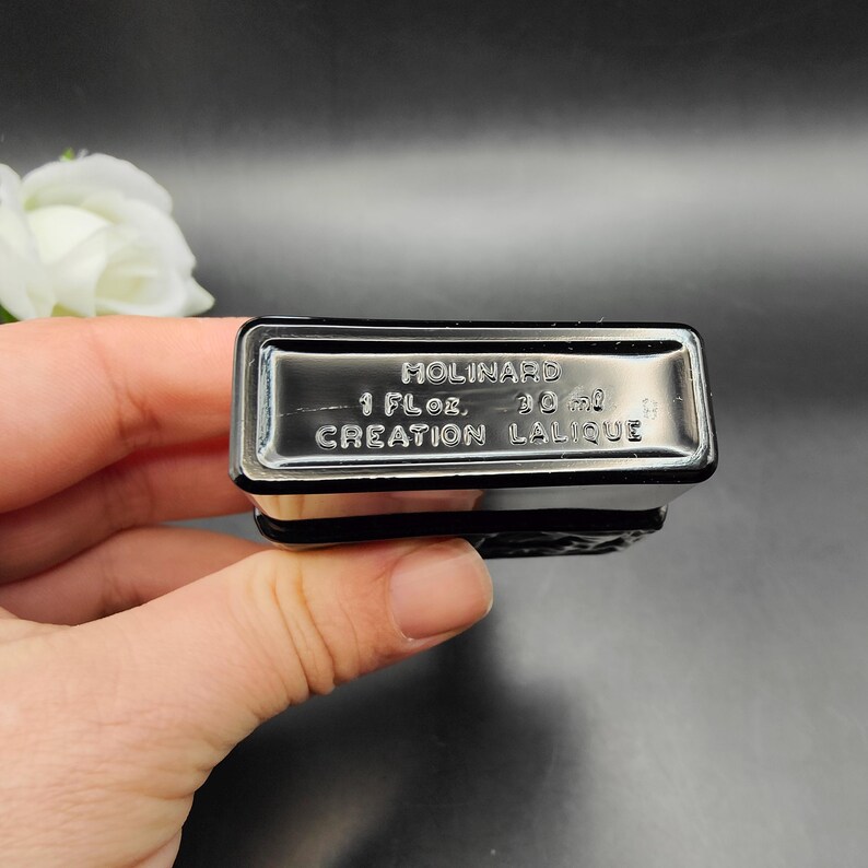 May include: A black rectangular perfume bottle with the text "Molinard" and "Creation Lalique". The bottle is labeled "1 fl oz" and "30 ml". The bottle is held in a person's hand.