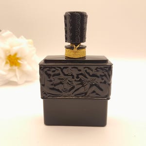 May include: A black perfume bottle with a decorative stopper and gold accents. The rectangular bottle has a detailed relief design. The stopper is cylindrical with a textured pattern. A white flower is in the background.