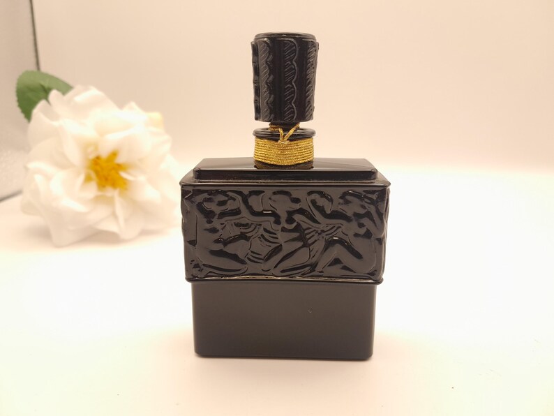 May include: A black perfume bottle with a decorative stopper and gold accents. The rectangular bottle has a detailed relief design. The stopper is cylindrical with a textured pattern. A white flower is in the background.