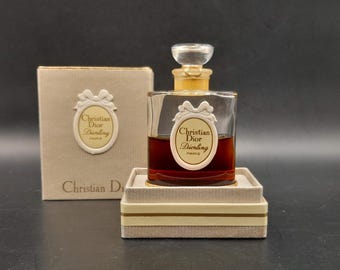 Könnte beinhalten: Eine Vintage-Flasche Christian Dior Diorling Parfum in einer klaren Glasflasche mit goldener Kappe und weißem Etikett. Die Flasche steht auf einer weißen Schachtel mit goldenen Verzierungen. Die Schachtel trägt den Schriftzug "Christian Dior".