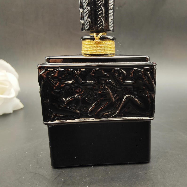 May include: A black perfume bottle with a decorative stopper. The bottle has a rectangular base and is adorned with a relief sculpture of figures. The stopper is black with a gold-colored band.