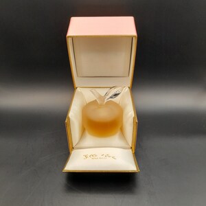 May include: A pink box with a gold trim is open, revealing a clear glass apple-shaped bottle of perfume. The bottle is filled with a light yellow liquid. The box is lined with white satin and has the words "L'Air du Temps Nina Ricci" printed on the inside.