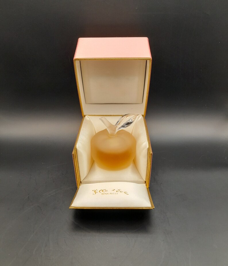 May include: A pink box with a gold trim is open, revealing a clear glass apple-shaped bottle of perfume. The bottle is filled with a light yellow liquid. The box is lined with white satin and has the words "L'Air du Temps Nina Ricci" printed on the inside.