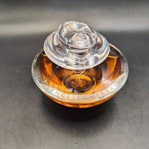 Puede incluir: Un frasco de perfume de vidrio transparente con una tapa dorada y una base dorada. El frasco est&aacute; etiquetado con "Silence Guerlain".
