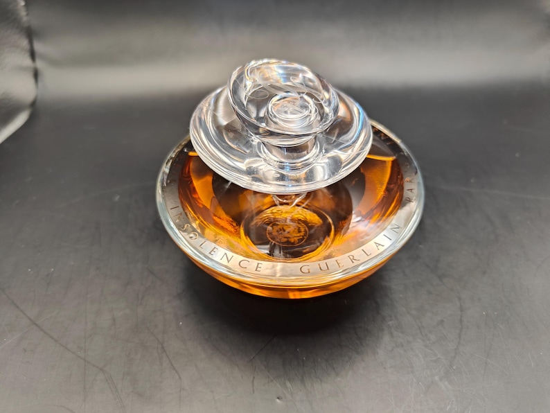 Puede incluir: Un frasco de perfume de vidrio transparente con una tapa dorada y una base dorada. El frasco est&aacute; etiquetado con "Silence Guerlain".