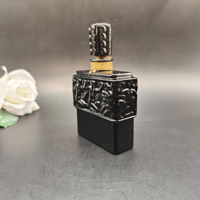 May include: A black perfume bottle with a rectangular base and a decorative top. The top features carved figures and a patterned stopper. The bottle is accented with gold-colored thread.
