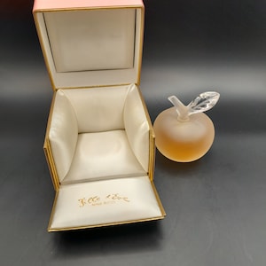 May include: A pink box with a gold trim and a white satin interior. The box is open and the interior is visible. The box is labeled "Fille d'Eve Nina Ricci". A clear glass bottle shaped like an apple with a gold lid is sitting on a black surface.