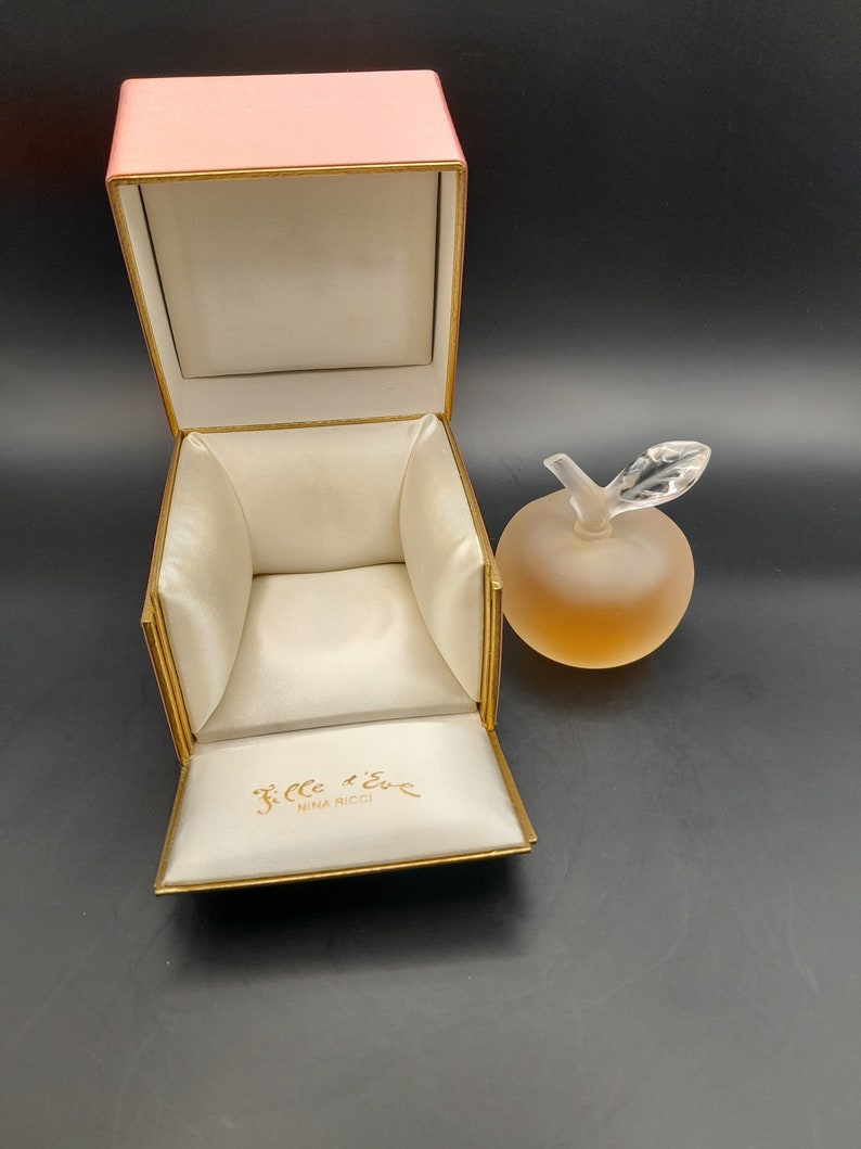 May include: A pink box with a gold trim and a white satin interior. The box is open and the interior is visible. The box is labeled "Fille d'Eve Nina Ricci". A clear glass bottle shaped like an apple with a gold lid is sitting on a black surface.