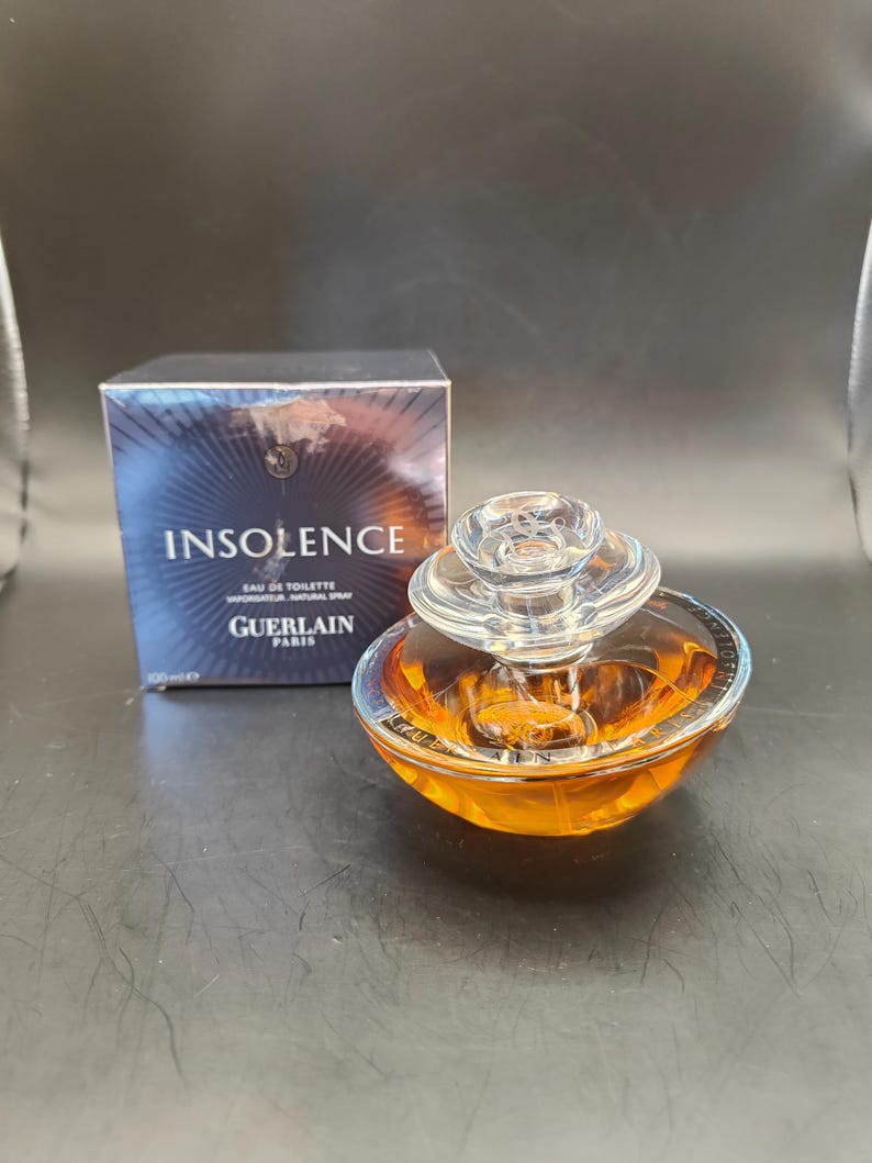 Puede incluir: Un frasco de perfume Guerlain Insolence Eau de Toilette de vidrio transparente. El frasco tiene forma de esfera con una tapa plateada. El frasco est&aacute; sobre una superficie oscura. La caja del perfume est&aacute; en el fondo. La caja es azul y plateada con las palabras "Insolence" y "Guerlain Paris" impresas en ella.