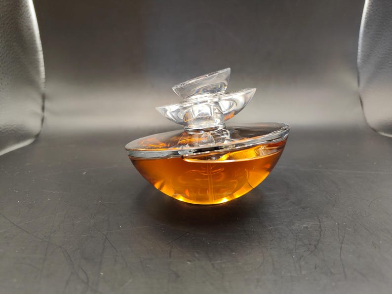 Puede incluir: Un frasco de perfume de vidrio transparente con un l&iacute;quido dorado en su interior. El frasco tiene un dise&ntilde;o &uacute;nico y en capas con una tapa plateada.